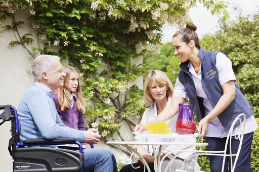 Nurse bringing drinks to her client who is sat in his wheelchair in the garden with his wife and daughter